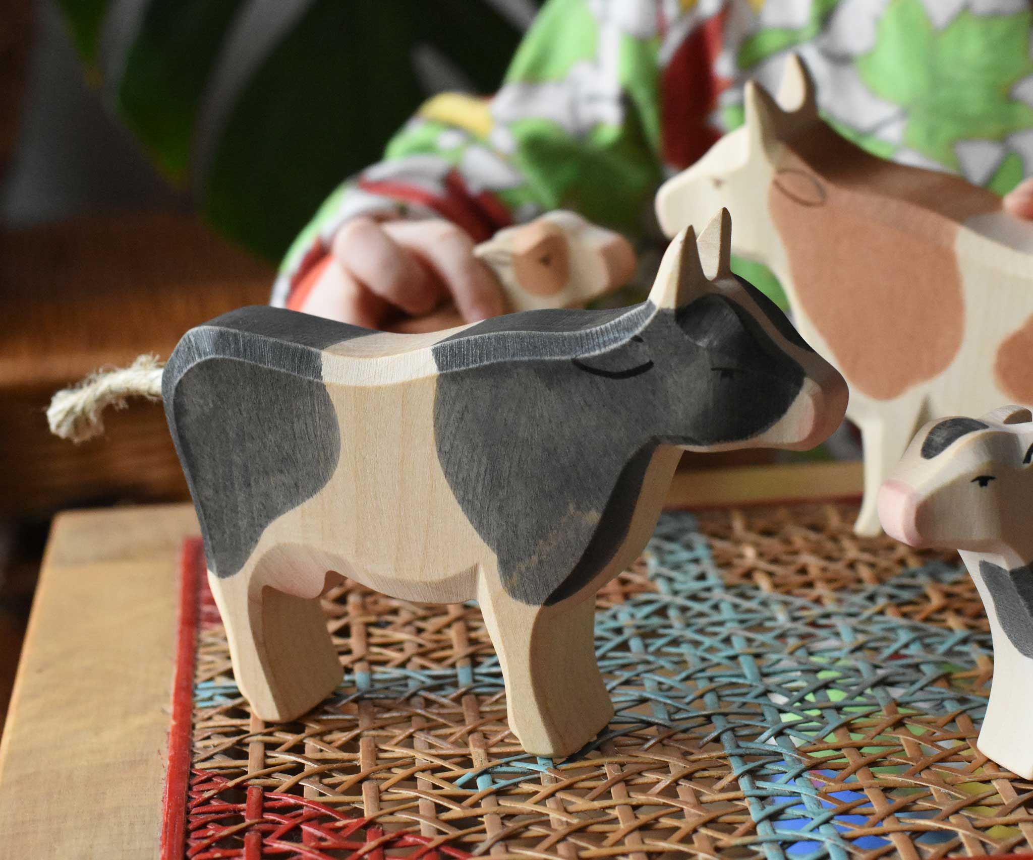 Ostheimer black and white cow wooden toy figure placed on a rattan stool in the middle of a simple small world play scene featuring cows and calves.