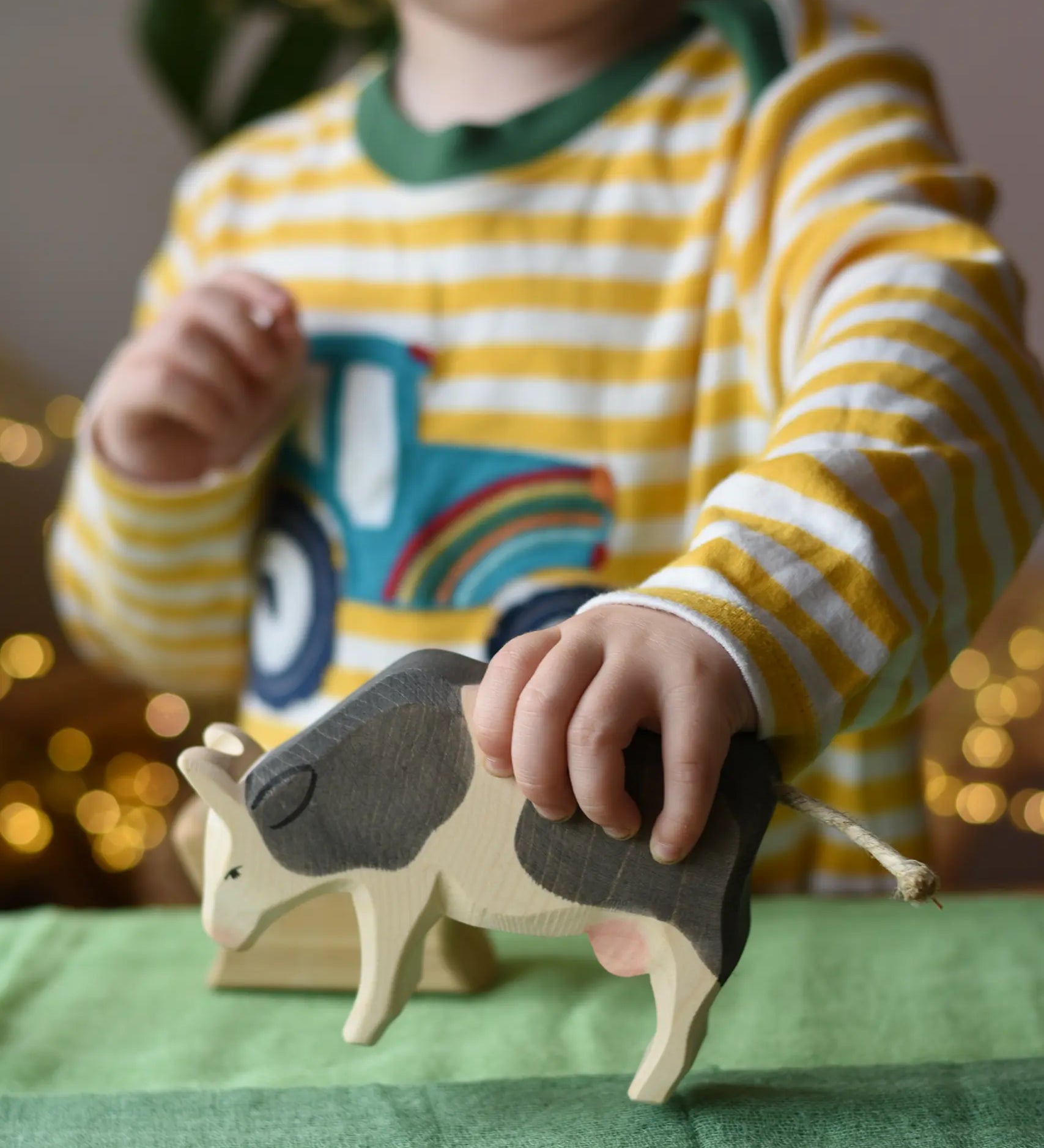 Ostheimer black and white cow eating wooden toy figure in a child's hand being lifted off the green cotton play cloths placed underneath.