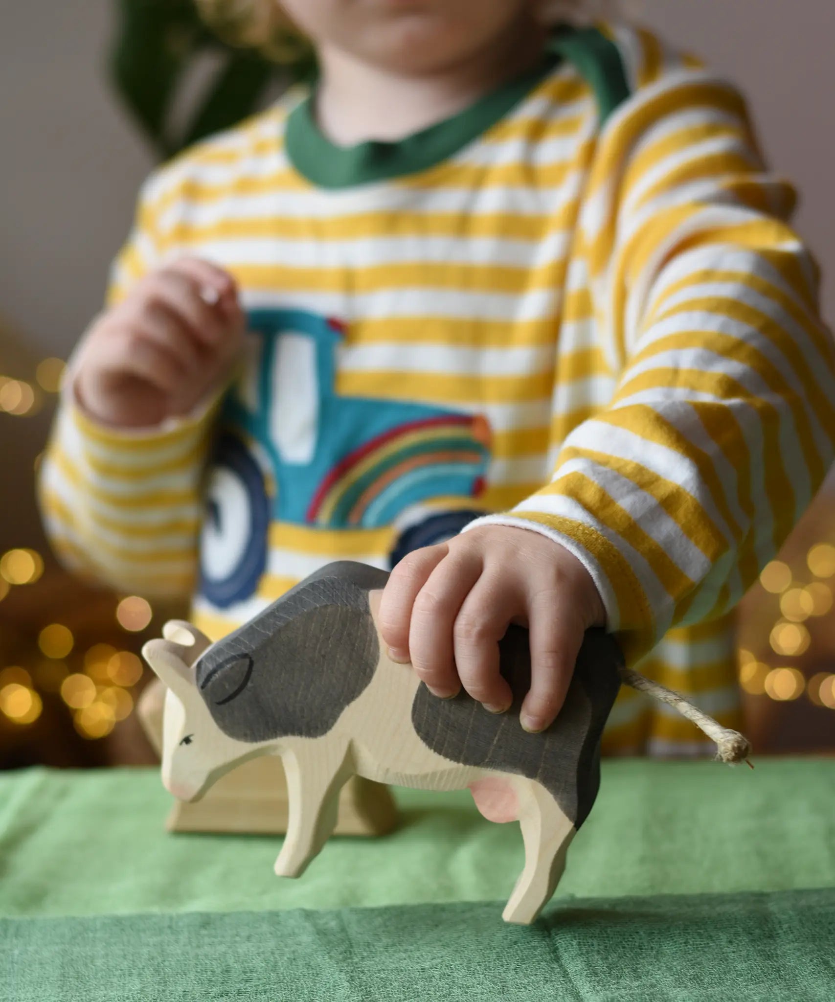 Ostheimer black and white cow eating wooden toy figure in a child's hand being lifted off the green cotton play cloths placed underneath.