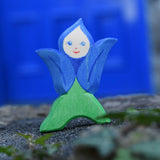 An Ostheimer hand made and hand painted wooden Gentian Flower Child figure toy placed on a stone wall. These flower child wooden figures are part of a wide range of expertly hand crafted wooden toys here at Babipur. 