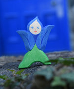 An Ostheimer hand made and hand painted wooden Gentian Flower Child figure toy placed on a stone wall. These flower child wooden figures are part of a wide range of expertly hand crafted wooden toys here at Babipur. 