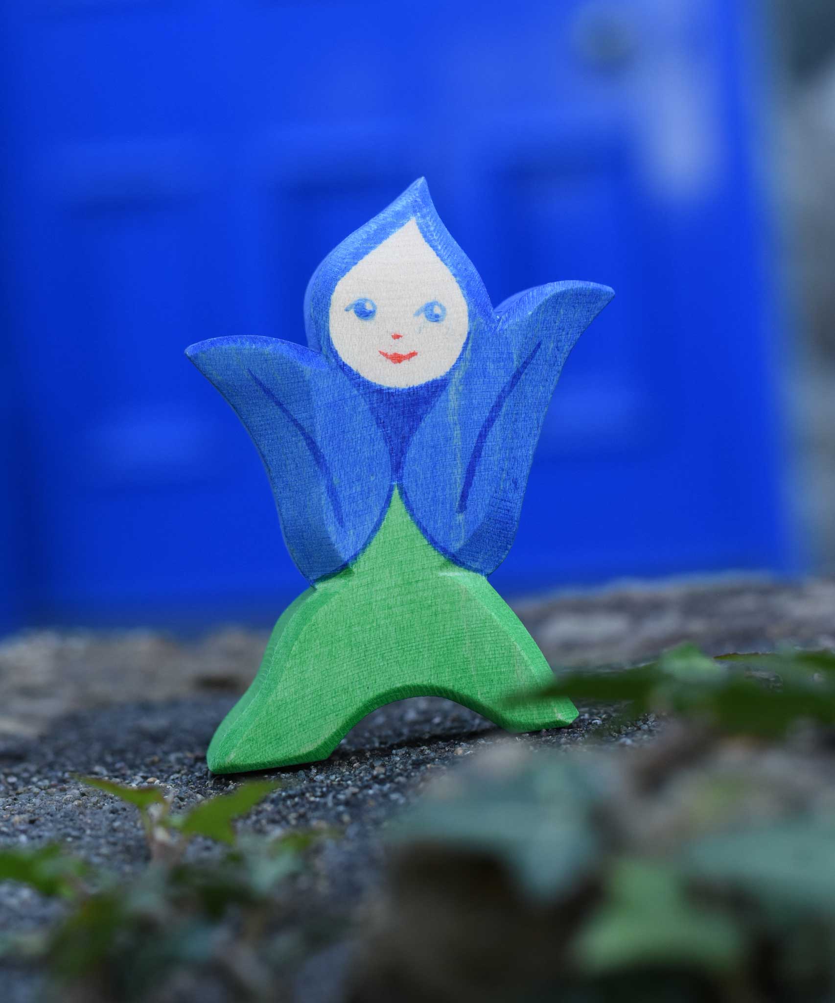 An Ostheimer hand made and hand painted wooden Gentian Flower Child figure toy placed on a stone wall. These flower child wooden figures are part of a wide range of expertly hand crafted wooden toys here at Babipur. 