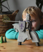 The Ostheimer blue riding wooden knight on a grey horse toy figure placed on a wooden surface with a child looking on in the background. 