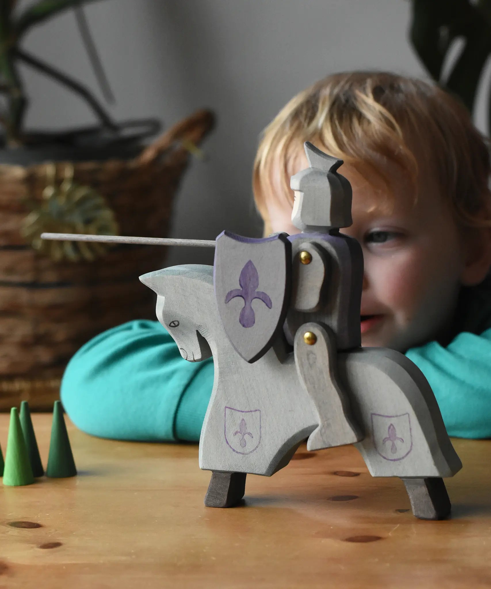 The Ostheimer blue riding wooden knight on a grey horse toy figure placed on a wooden surface with a child looking on in the background. 