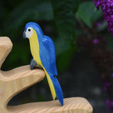 Ostheimer Blue and yellow parrot figurine on a wooden perch with a blurred purple flower background