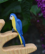 Ostheimer Blue and yellow parrot figurine on a wooden perch with a blurred purple flower background
