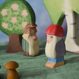 A side profile view of Bodo the dwarf laced in a small world play scene. The wooden toy figure has a long white beard, red hat, purple coat, blue trousers and brown boots. There are other wooden toy in the background including trees and a fir tree dwarf. These wooden figures are part of a large range of expertly hand crafted, Waldorf inspired, wooden toys here at Babipur. 