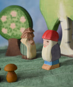 A side profile view of Bodo the dwarf laced in a small world play scene. The wooden toy figure has a long white beard, red hat, purple coat, blue trousers and brown boots. There are other wooden toy in the background including trees and a fir tree dwarf. These wooden figures are part of a large range of expertly hand crafted, Waldorf inspired, wooden toys here at Babipur. 