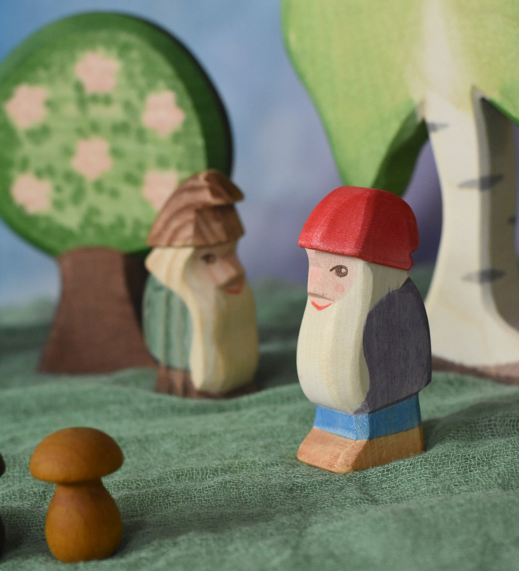 A side profile view of Bodo the dwarf laced in a small world play scene. The wooden toy figure has a long white beard, red hat, purple coat, blue trousers and brown boots. There are other wooden toy in the background including trees and a fir tree dwarf. These wooden figures are part of a large range of expertly hand crafted, Waldorf inspired, wooden toys here at Babipur. 