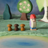 An Ostheimer Bodo wooden dwarf figure placed at the centre of a small world play scene next to three Grapat wooden mushroom mandala pieces. The Ostherimer pond scene piece has been placed in front of the dwarf. Ostheimer trees can be seen in the background. These wooden figures are part of a large range of expertly hand crafted, Waldorf inspired, wooden toys here at Babipur. 