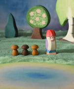 An Ostheimer Bodo wooden dwarf figure placed at the centre of a small world play scene next to three Grapat wooden mushroom mandala pieces. The Ostherimer pond scene piece has been placed in front of the dwarf. Ostheimer trees can be seen in the background. These wooden figures are part of a large range of expertly hand crafted, Waldorf inspired, wooden toys here at Babipur. 