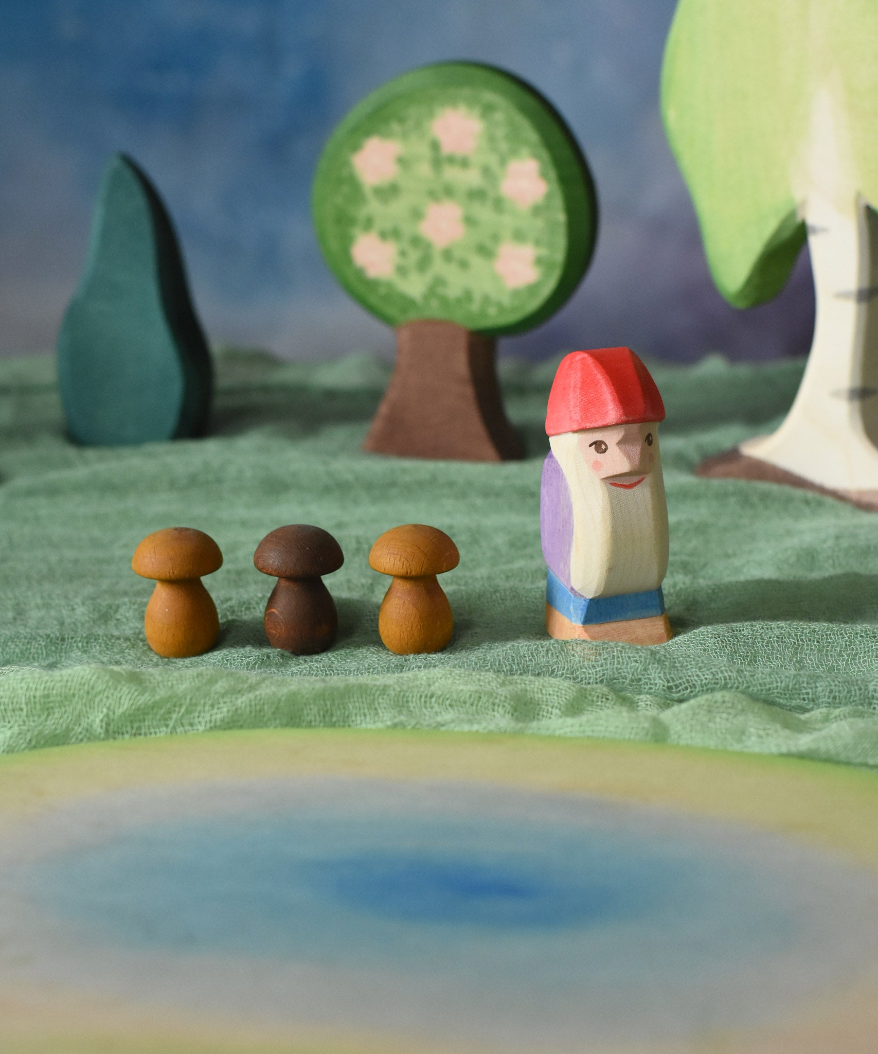 An Ostheimer Bodo wooden dwarf figure placed at the centre of a small world play scene next to three Grapat wooden mushroom mandala pieces. The Ostherimer pond scene piece has been placed in front of the dwarf. Ostheimer trees can be seen in the background. These wooden figures are part of a large range of expertly hand crafted, Waldorf inspired, wooden toys here at Babipur. 