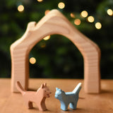 An Ostheimer Bremer dog and cat wooden toy figure placed on a wooden surface with a house shaped wooden piece in the background.  Ostheimer's range of hand crafted, heirloom quality wooden toys are available to buy at Babipur. 