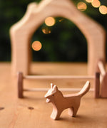 An Ostheimer Bremer dog wooden toy figure placed on a wooden surface with a house shaped wooden piece in the background.  Ostheimer's range of hand crafted, heirloom quality wooden toys are available to buy at Babipur. 