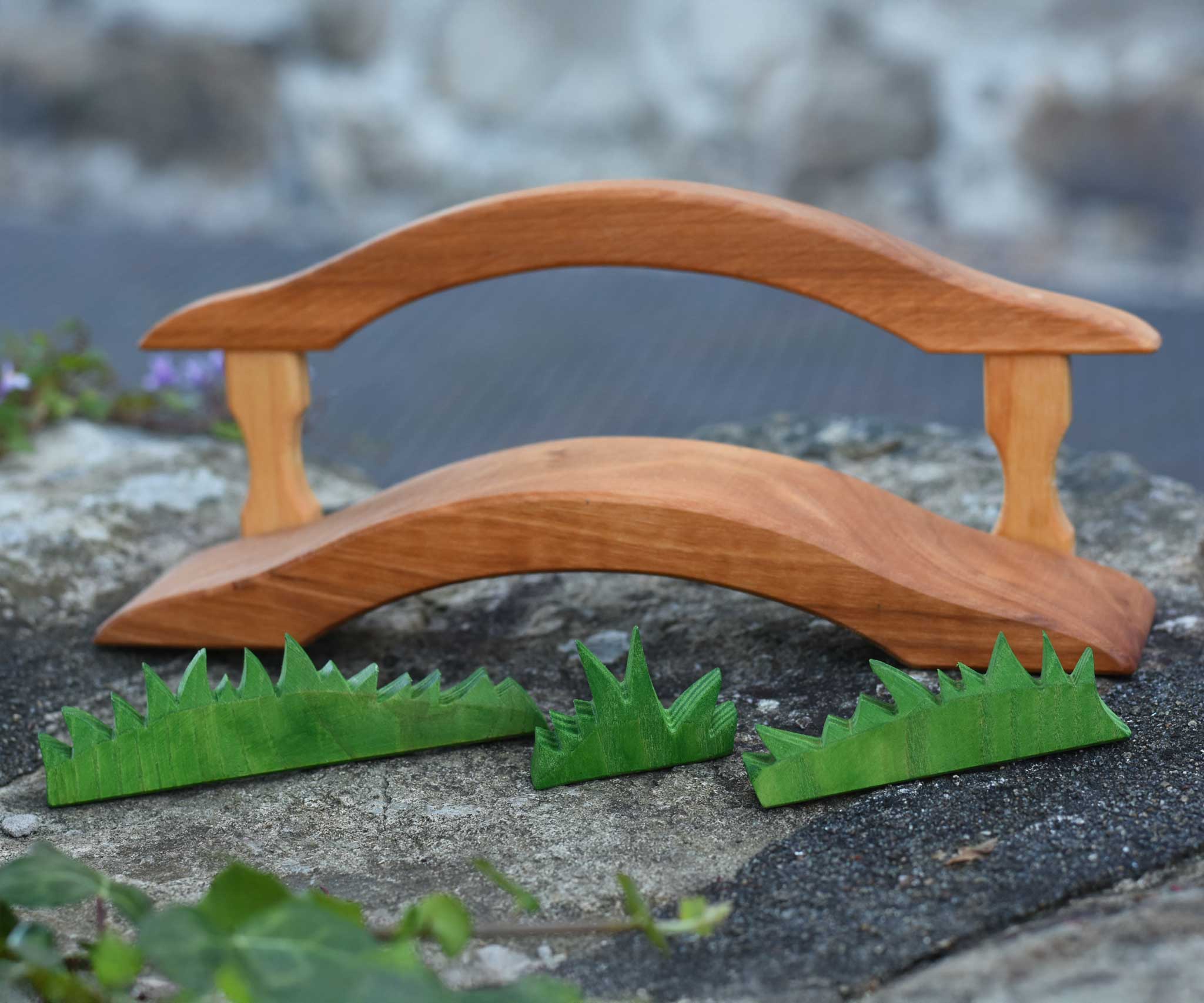 An Ostheimer wooden bridge with railing placed on a stone wall outdoor. Ostheimer grass pieces have been placed in front of the bridge.