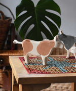 Ostheimer brown standing cow figure placed on a rattan stool in a simple small world play scene with other cows and calves in the background. 