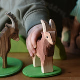 Child's hand holding the Ostheimer brown male goat wooden toy figure on a small green meadow plate. 