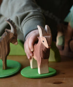 Child's hand holding the Ostheimer brown male goat wooden toy figure on a small green meadow plate. 
