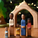 A family of Ostheimer figures with brown skin. There is a mother, father, son, daughter and toddler figure. The figures are all lined up and facing facing the camera showing their fronts. The wooden house shaped piece from the Ostheimer 4 piece house set can be seen in the background. These wooden figures are part of a wide range of expertly hand crafted wooden toys here at Babipur.