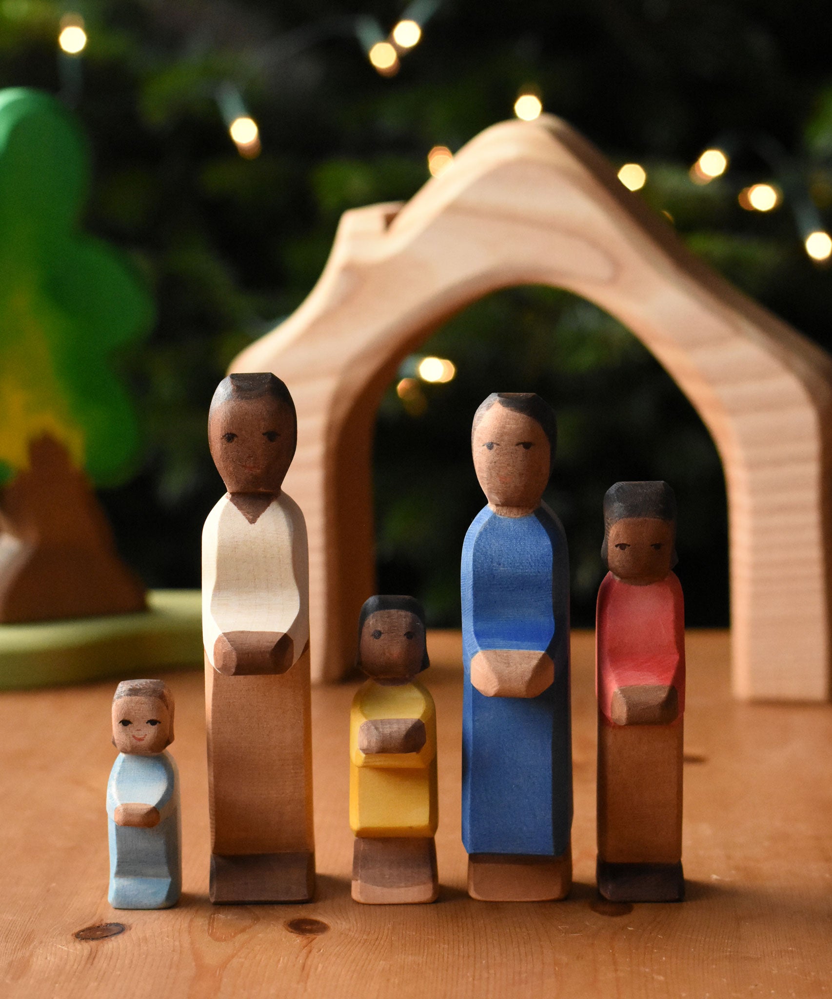 A family of Ostheimer figures with brown skin. There is a mother, father, son, daughter and toddler figure. The figures are all lined up and facing facing the camera showing their fronts. The wooden house shaped piece from the Ostheimer 4 piece house set can be seen in the background. These wooden figures are part of a wide range of expertly hand crafted wooden toys here at Babipur.