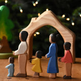 A family of Ostheimer figures with brown skin. There is a mother, father, son, daughter and toddler figure. The figures are all lined up and facing to the left showing their side profiles. The wooden house shaped piece from the Ostheimer 4 piece house set can be seen in the background. These wooden figures are part of a wide range of expertly hand crafted wooden toys here at Babipur.