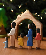 A family of Ostheimer figures with brown skin. There is a mother, father, son, daughter and toddler figure. The figures are all lined up and facing to the left showing their side profiles. The wooden house shaped piece from the Ostheimer 4 piece house set can be seen in the background. These wooden figures are part of a wide range of expertly hand crafted wooden toys here at Babipur.