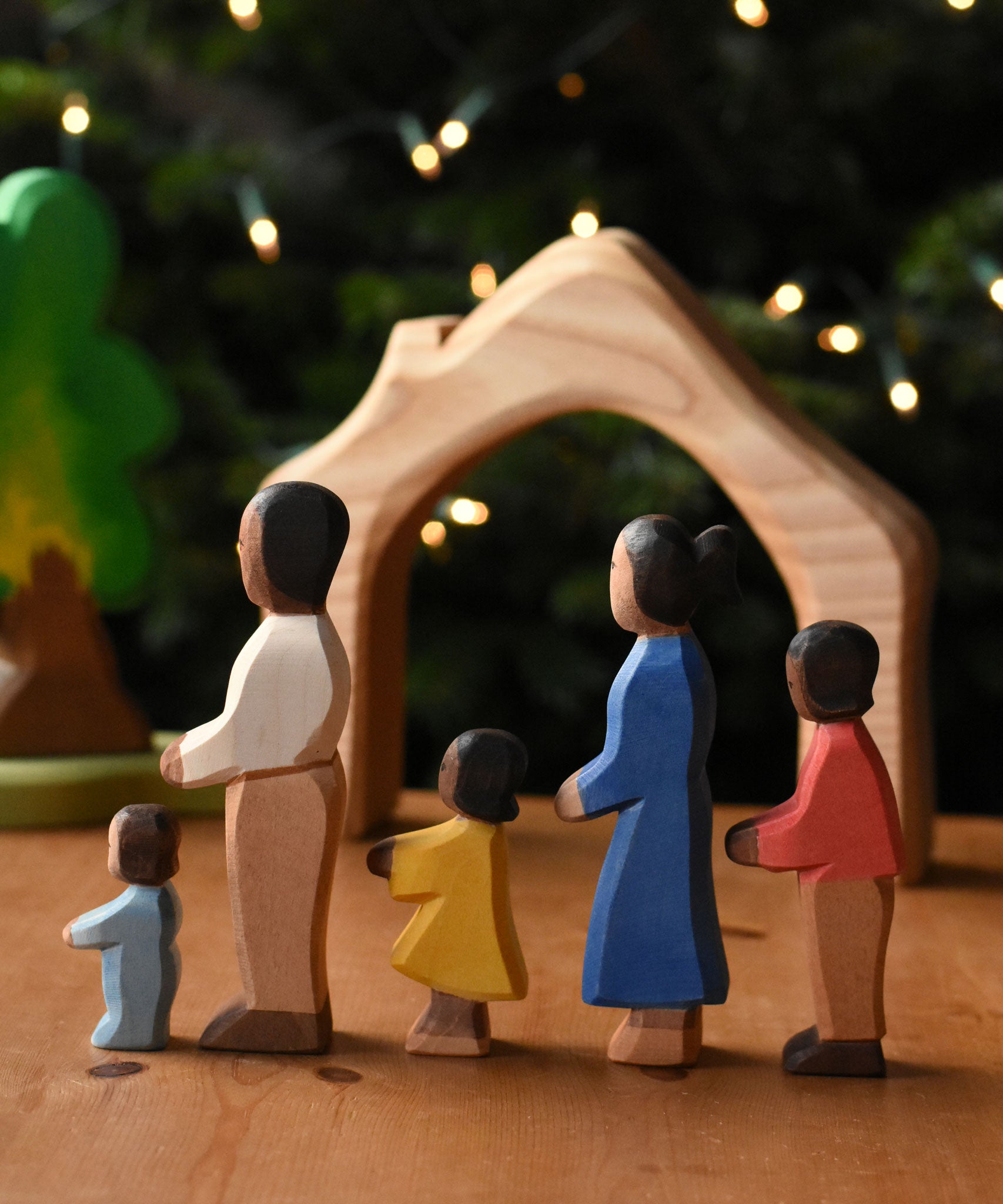 A family of Ostheimer figures with brown skin. There is a mother, father, son, daughter and toddler figure. The figures are all lined up and facing to the left showing their side profiles. The wooden house shaped piece from the Ostheimer 4 piece house set can be seen in the background. These wooden figures are part of a wide range of expertly hand crafted wooden toys here at Babipur.