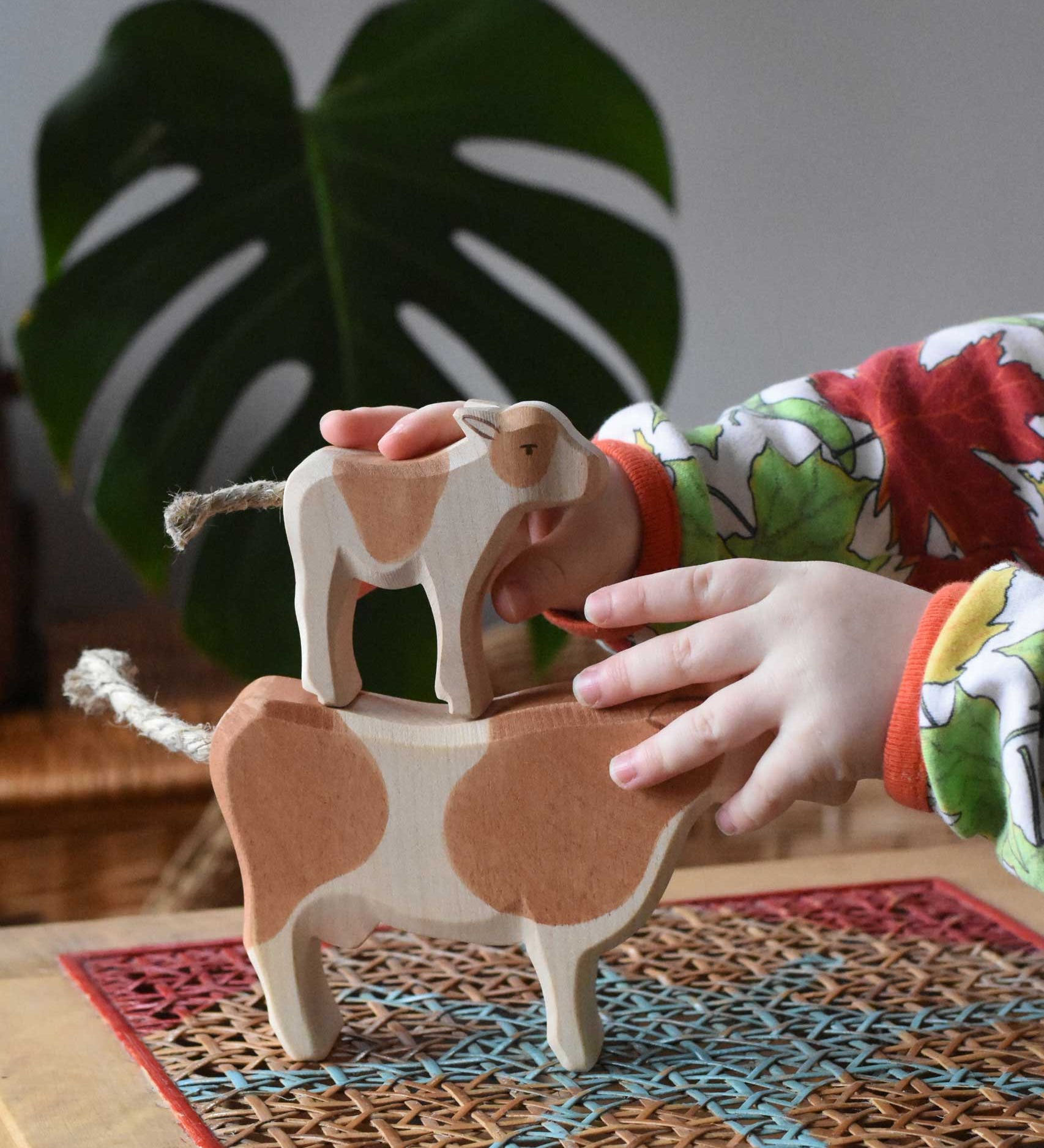 Child stacking a standing brown calf wooden toy figure on top of a brown cow figure.