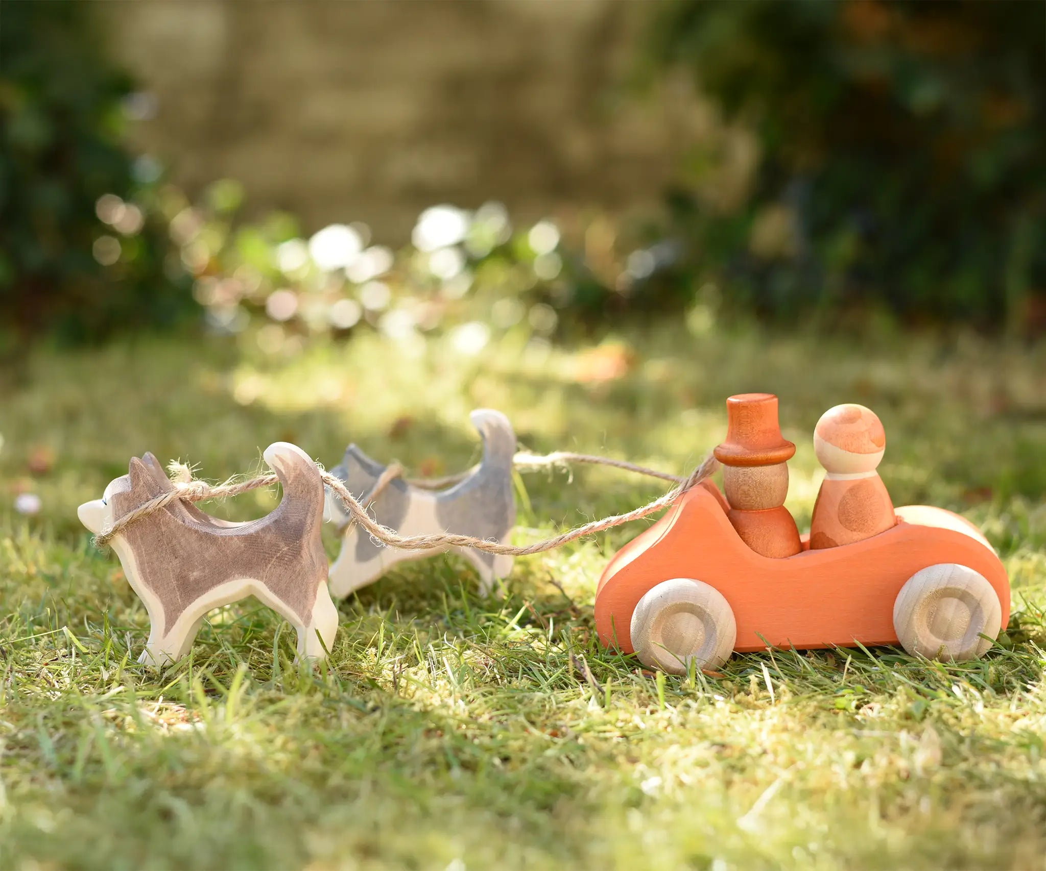 Ostheimer standing and running sled dogs tied by string to an orange Grimm's car with two Grapat peg dolls placed inside in the Babipur garden.