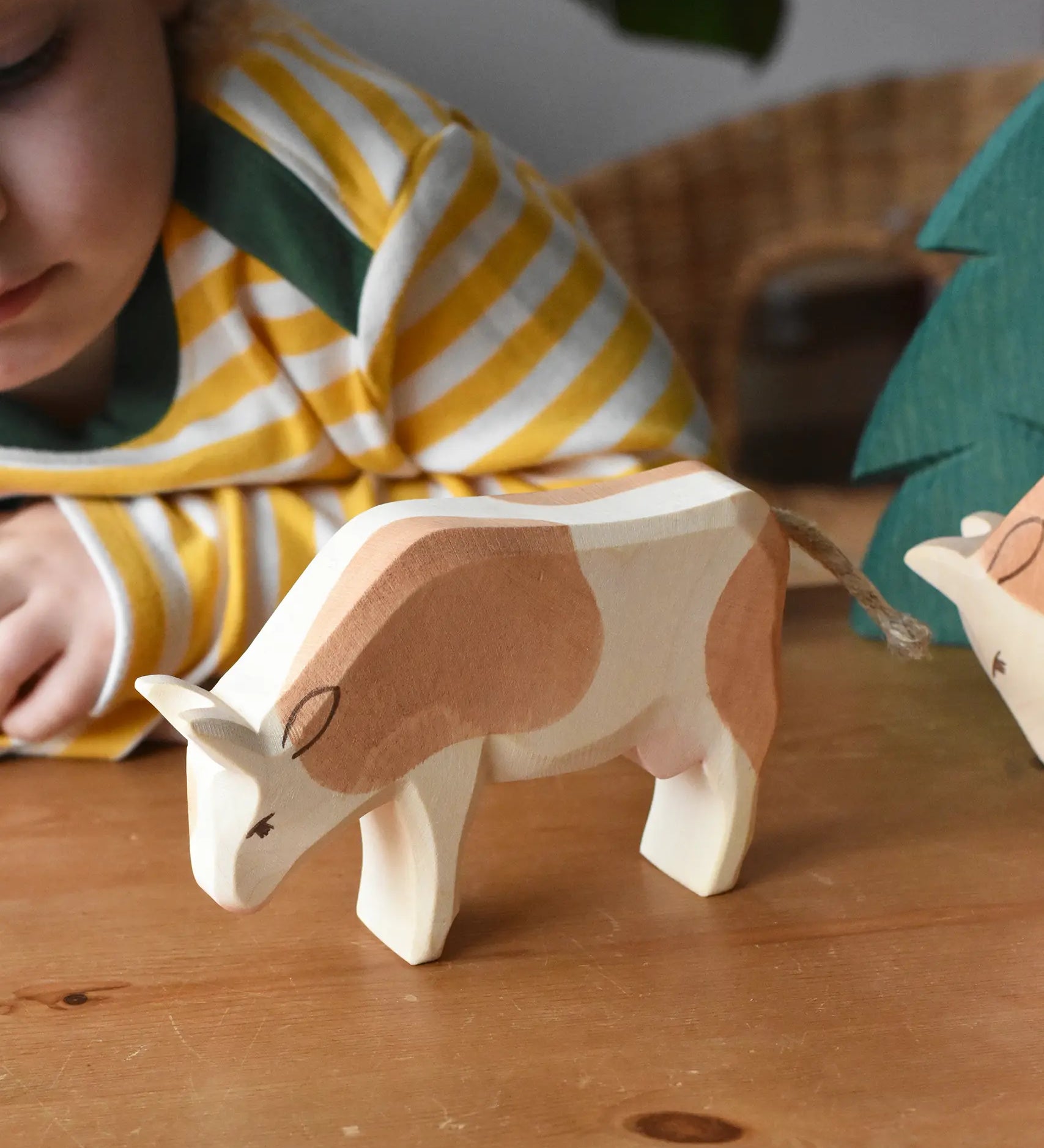 Ostheimer brown and white cow wooden toy figure placed on a wooden surface with a child in the background looking on.