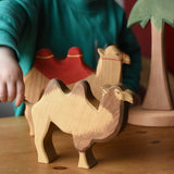 Ostheimer camel wooden toy figure placed in the front of a small world play scene. A child is holding the rope tail of the camel. 