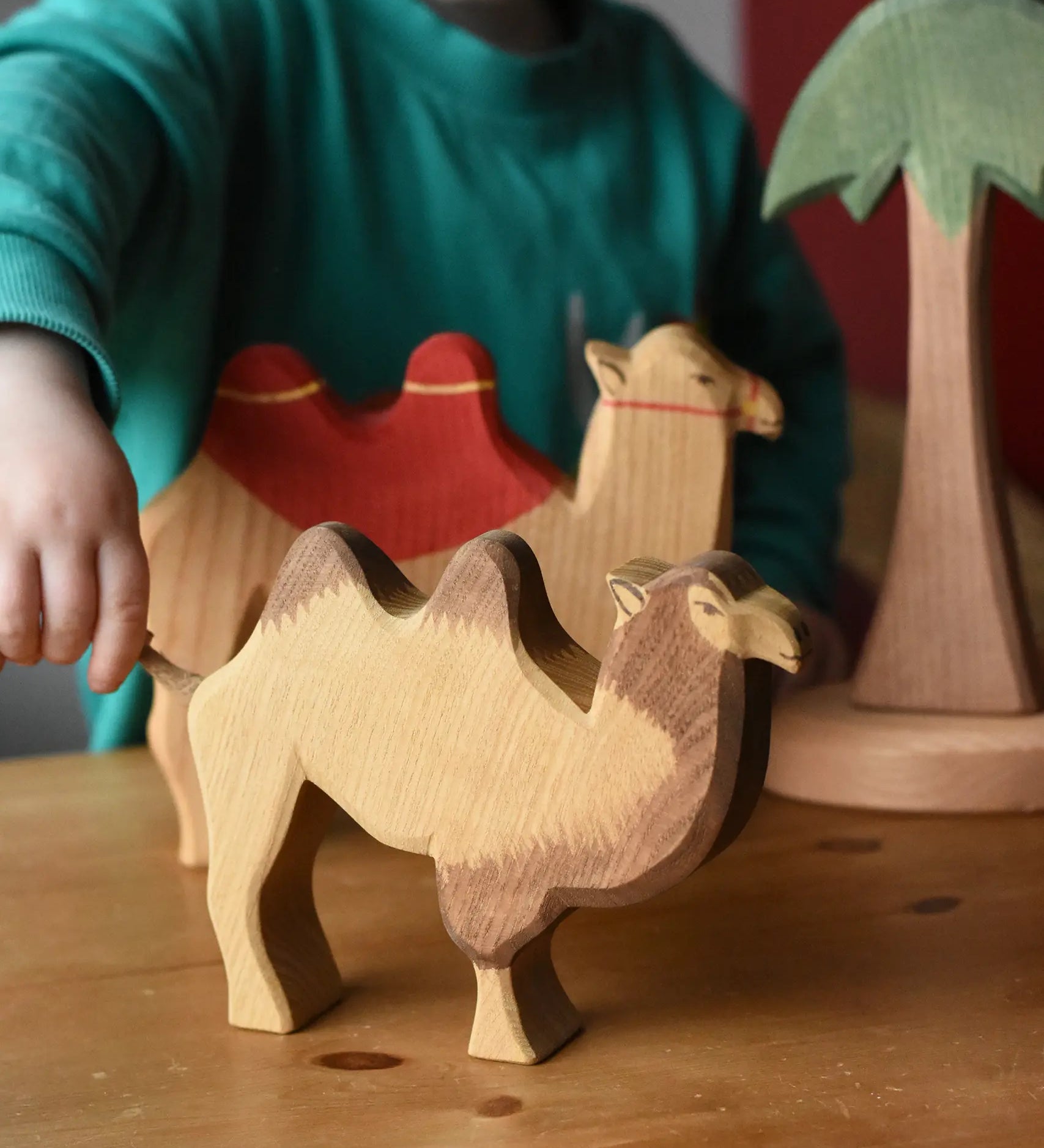 Ostheimer camel wooden toy figure placed in the front of a small world play scene. A child is holding the rope tail of the camel. 