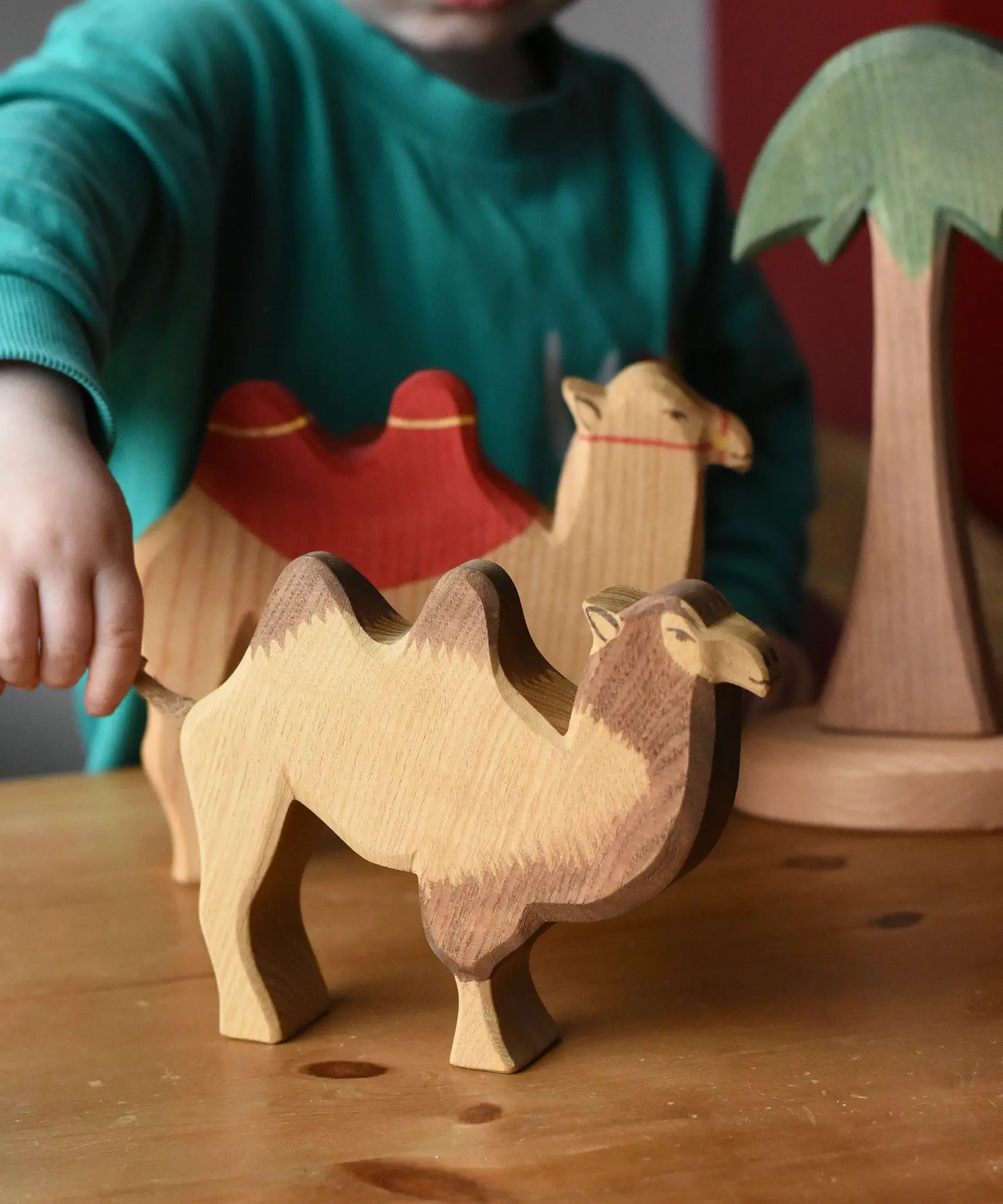 Ostheimer camel wooden toy figure placed in the front of a small world play scene. A child is holding the rope tail of the camel. 