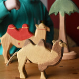 Ostheimer camel wooden toy figure placed on a wooden surface with another camel and tall palm tree in the background. 