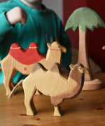 Ostheimer camel wooden toy figure placed on a wooden surface with another camel and tall palm tree in the background. 