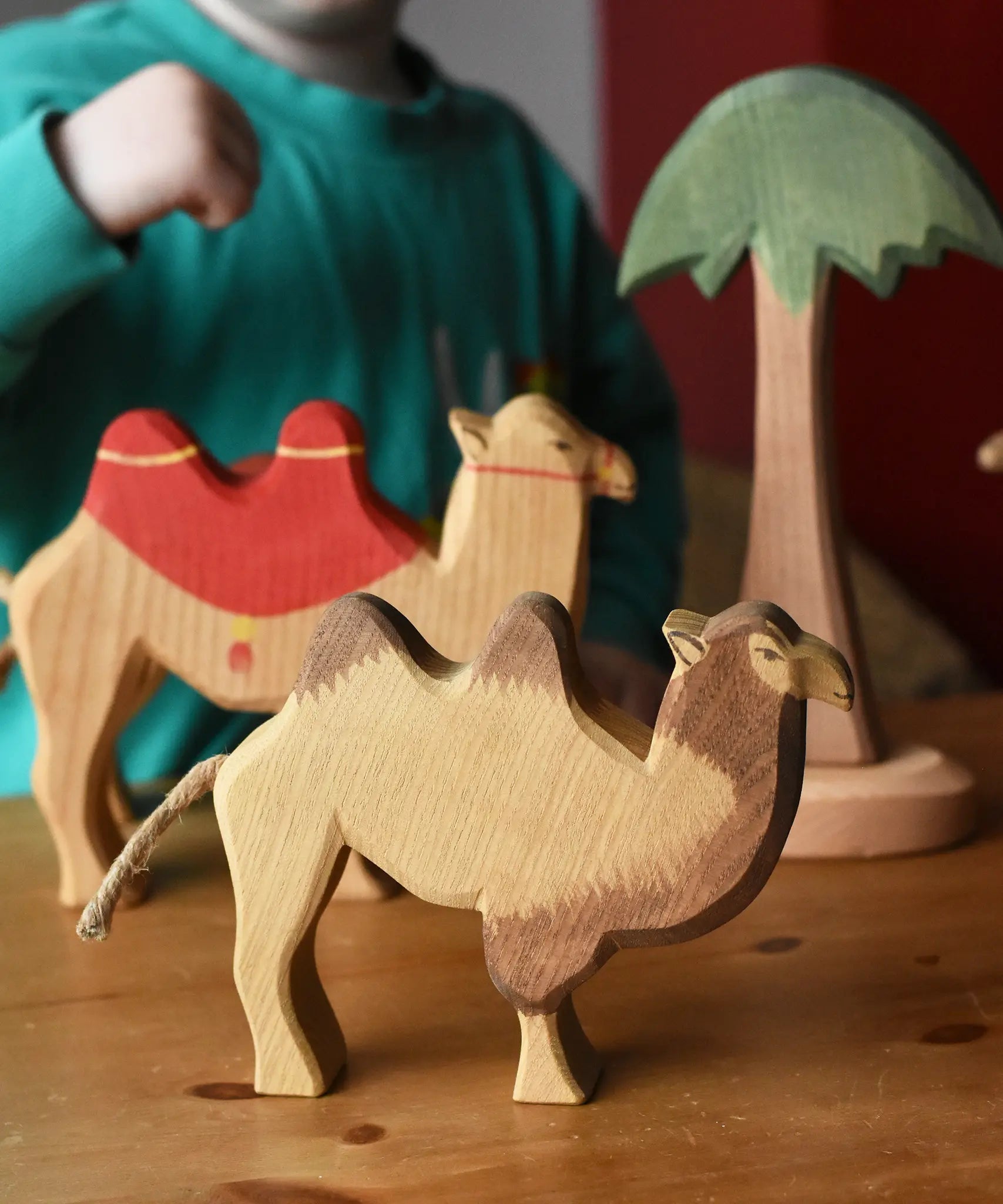 Ostheimer camel wooden toy figure placed on a wooden surface with another camel and tall palm tree in the background. 