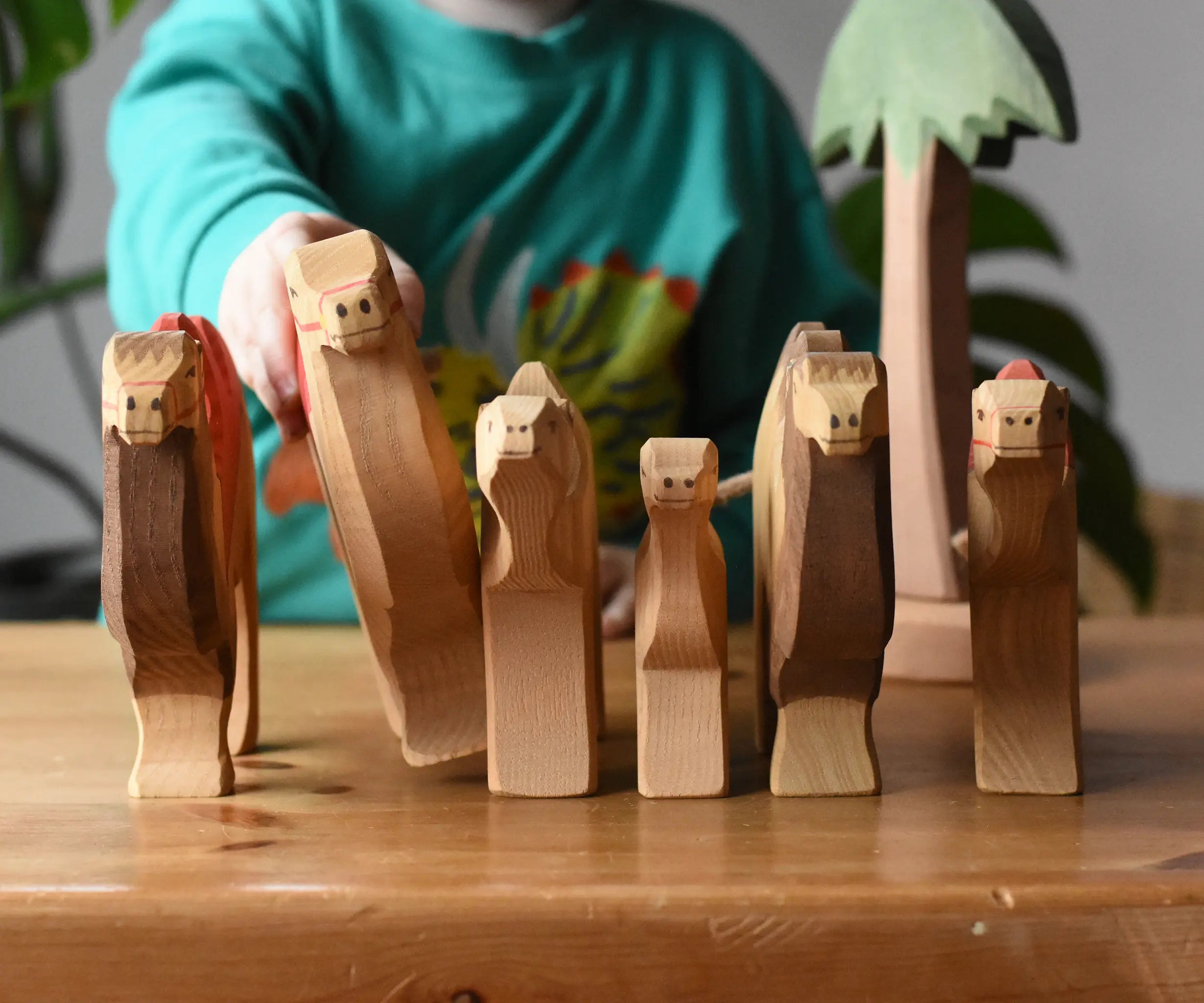 Six Ostheimer camel wooden toy figures lined up with a child holding one of them. Tall palm tree in the background.