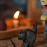 An Ostheimer hand made and hand painted wooden cat figure toy placed on a swooden surface. An Ostheimer witch and a lit candle on a Grimm's celebration ring can be seen in the background.These wooden figures are part of a wide range of expertly hand crafted wooden toys here at Babipur. 