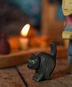 An Ostheimer hand made and hand painted wooden cat figure toy placed on a swooden surface. An Ostheimer witch and a lit candle on a Grimm's celebration ring can be seen in the background.These wooden figures are part of a wide range of expertly hand crafted wooden toys here at Babipur. 