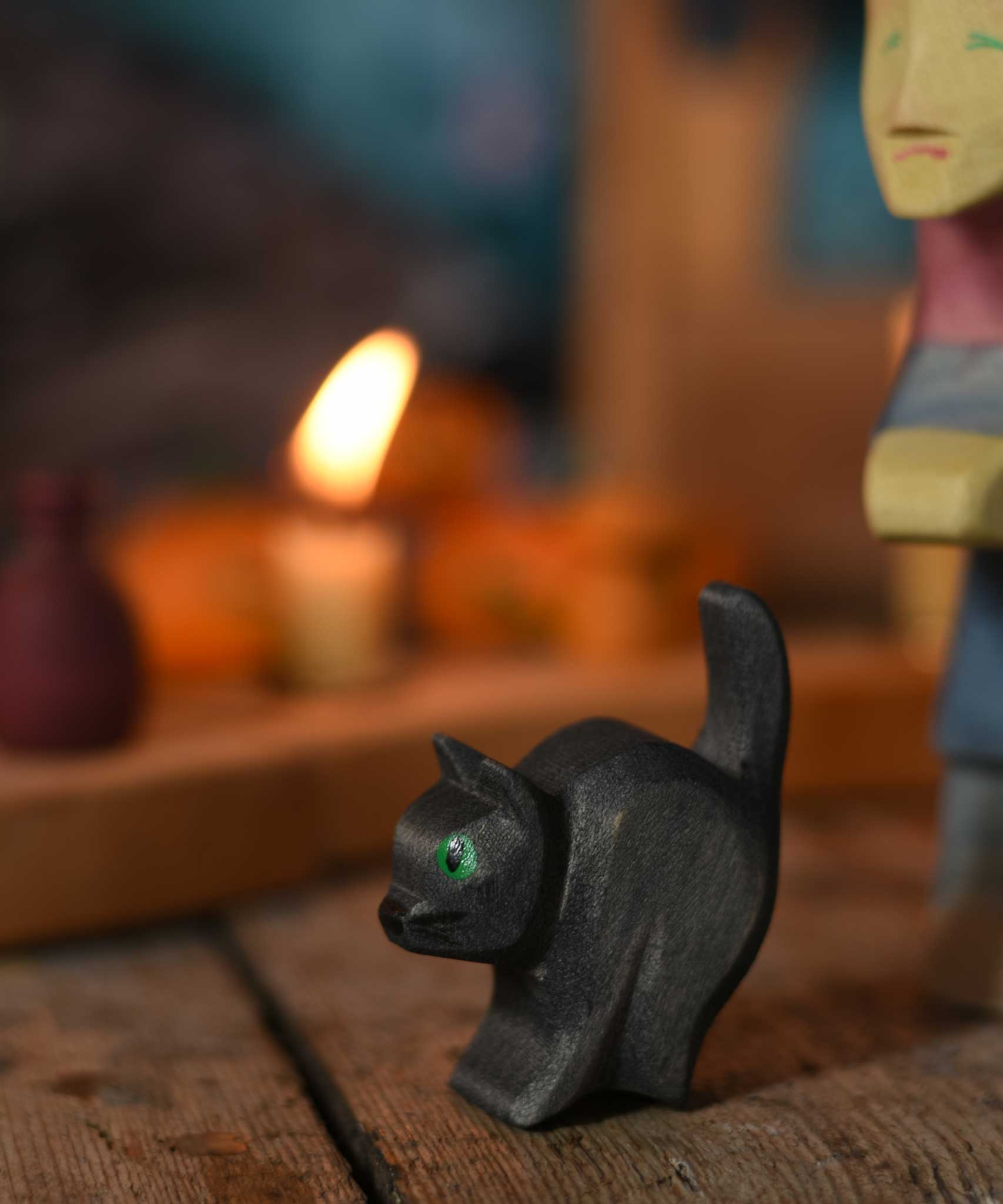 An Ostheimer hand made and hand painted wooden cat figure toy placed on a swooden surface. An Ostheimer witch and a lit candle on a Grimm's celebration ring can be seen in the background.These wooden figures are part of a wide range of expertly hand crafted wooden toys here at Babipur. 