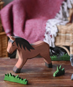 Ostheimer brown cold blood horse wooden toy figure placed in a simple small world play scene with wooden green grass scenery pieces. 