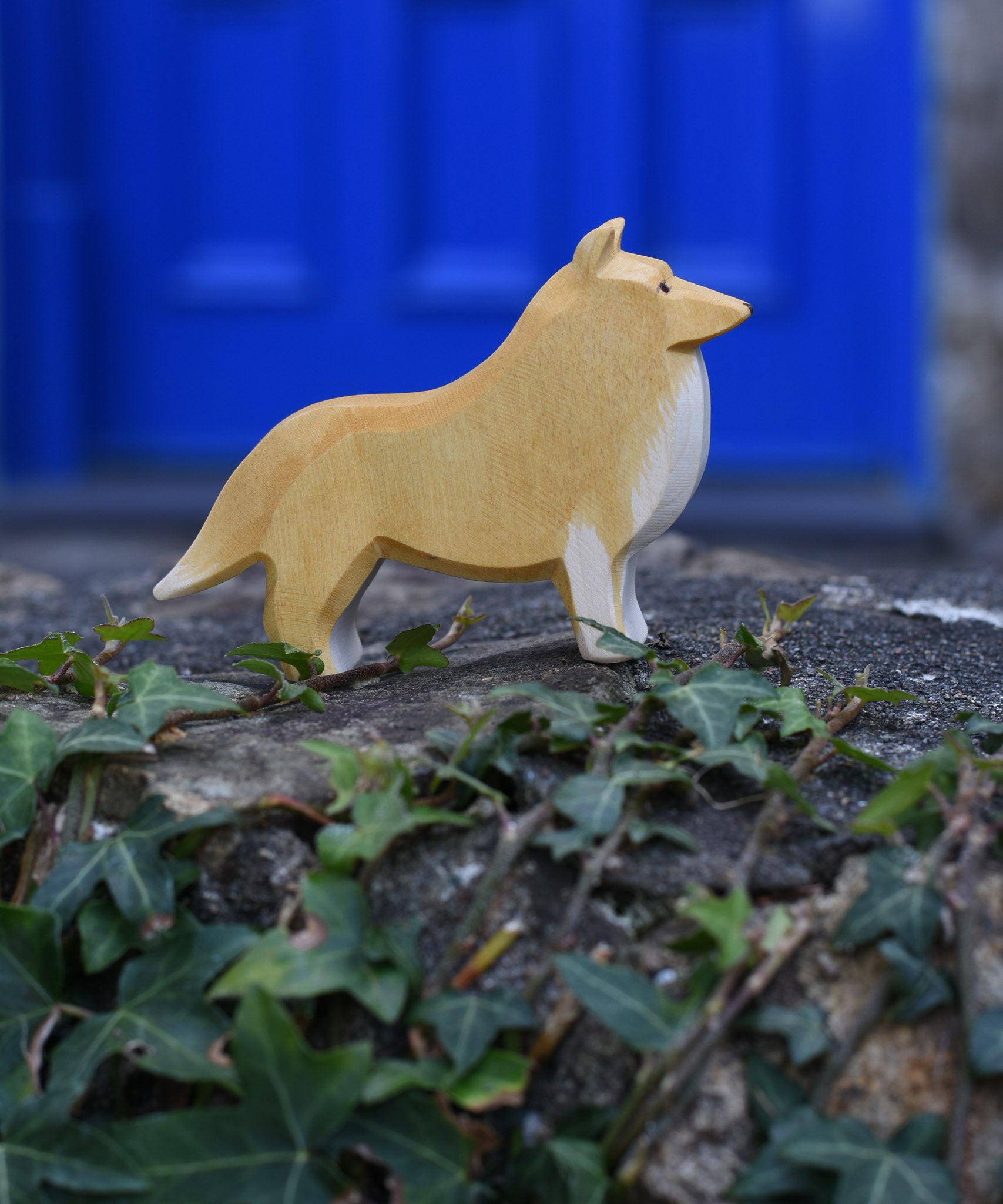 An Ostheimer Collie Dog Handmade Wooden Toy placed on a stone wall outdoors. These wooden figures are part of a large range of expertly hand crafted, Waldorf inspired, wooden toys here at Babipur.