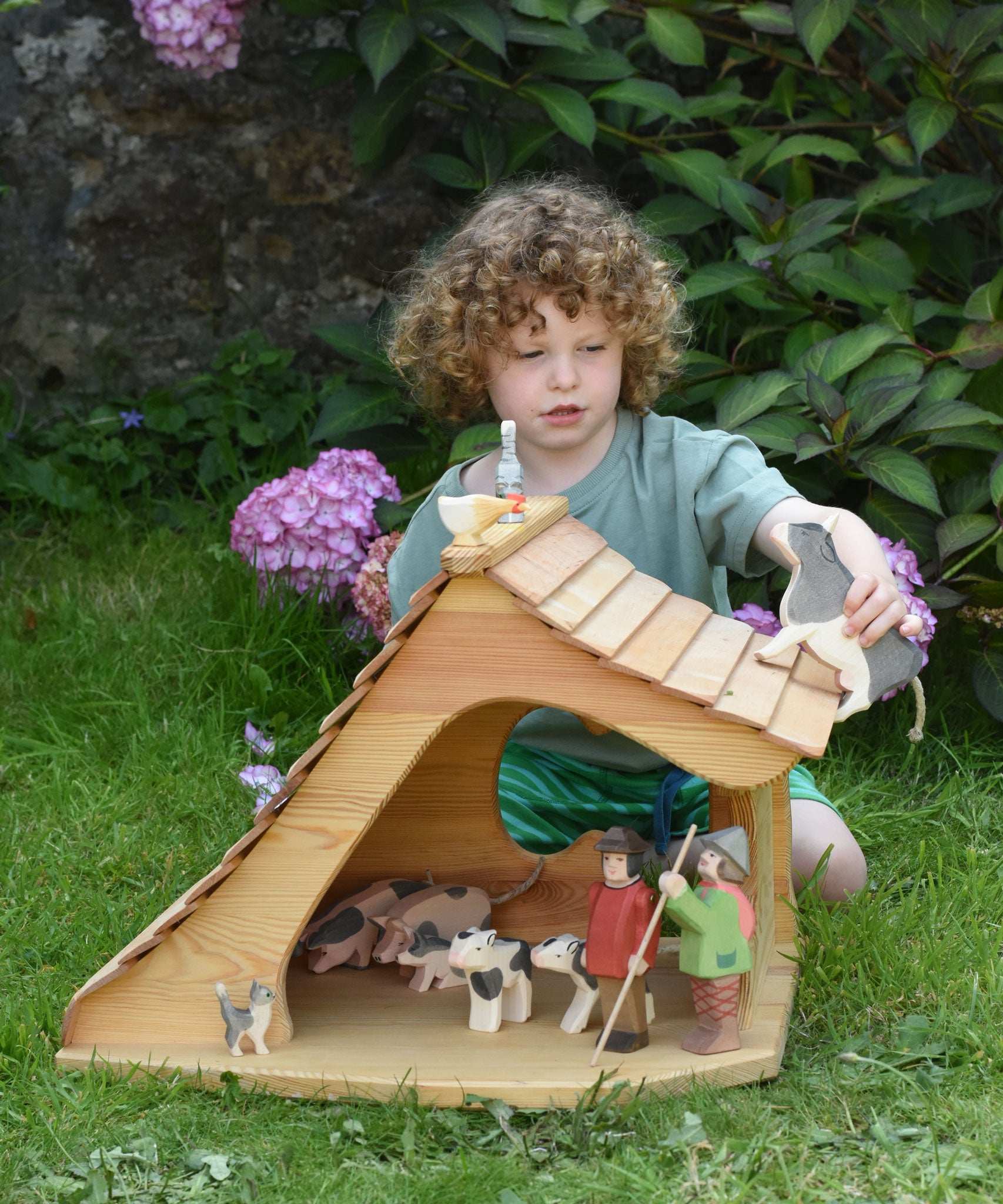 An Ostheimer wooden cottage dolls house filled with various Ostheimer animal and people figures. A child is holding a wooden cow figure, walking it across the roof of the cottage. 
These wooden figures are part of a large range of expertly hand crafted, Waldorf inspired, wooden toys here at Babipur. 