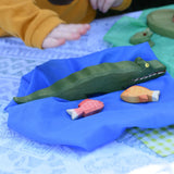 Ostheimer crocodile wooden toy figure placed next to red and yellow fish toy figures on a blue Laura's cotton play cloth.