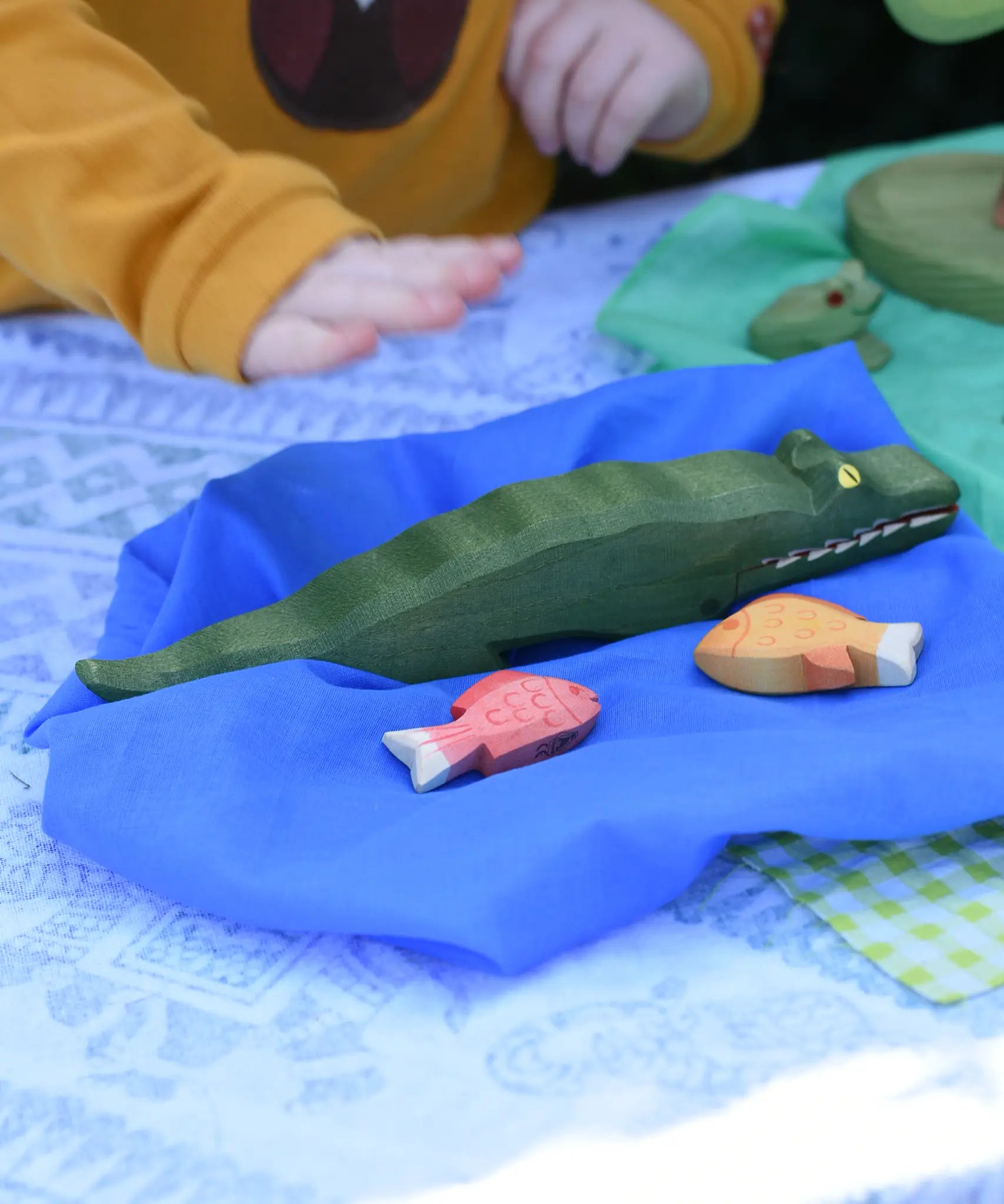 Ostheimer crocodile wooden toy figure placed next to red and yellow fish toy figures on a blue Laura's cotton play cloth.