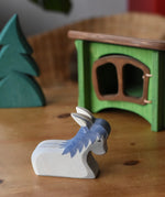Ostheimer donkey laying down wooden toy figure placed in a simple small world play scene with a small spruce tree in the background and a green hutch. 