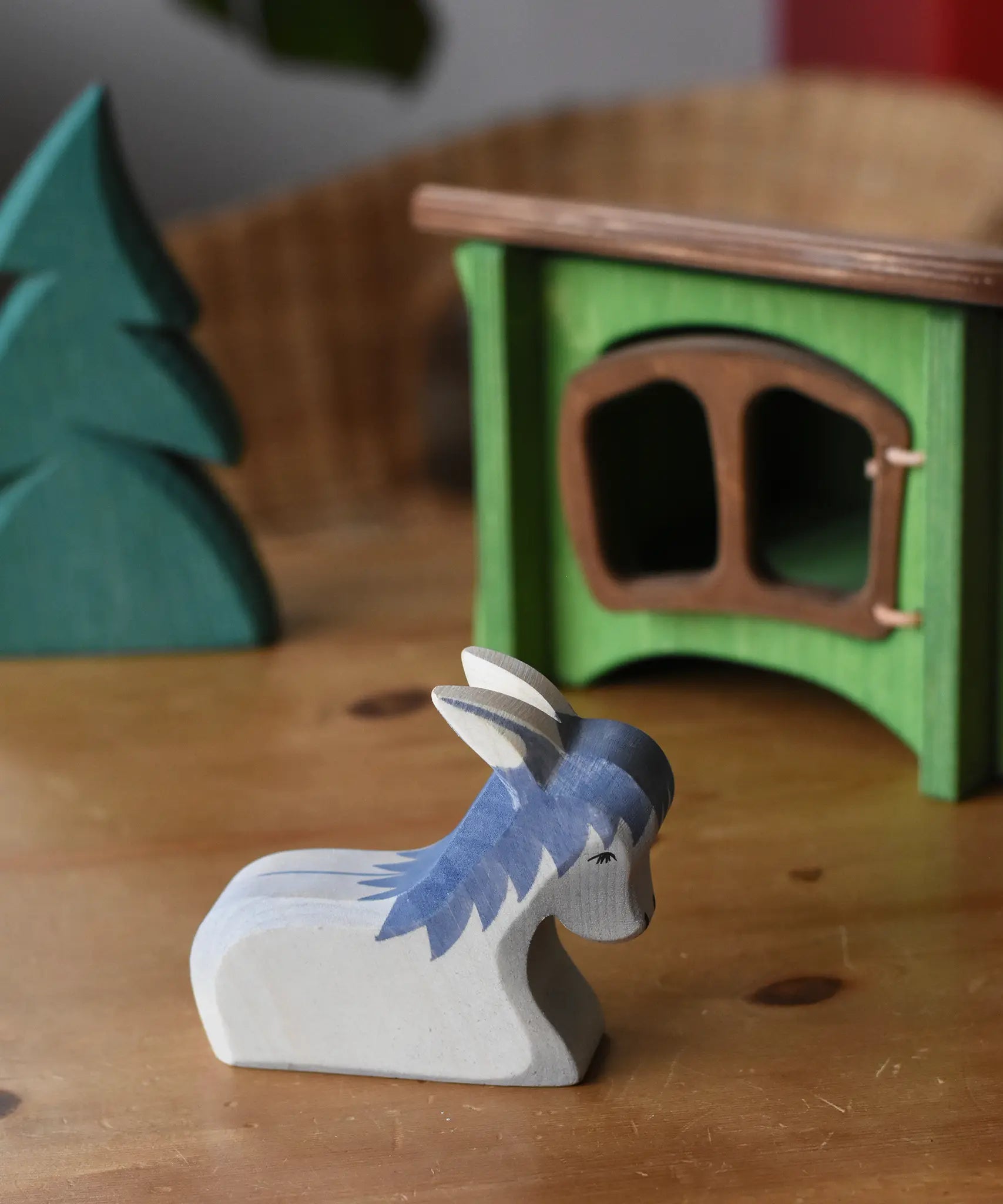 Ostheimer donkey laying down wooden toy figure placed in a simple small world play scene with a small spruce tree in the background and a green hutch. 