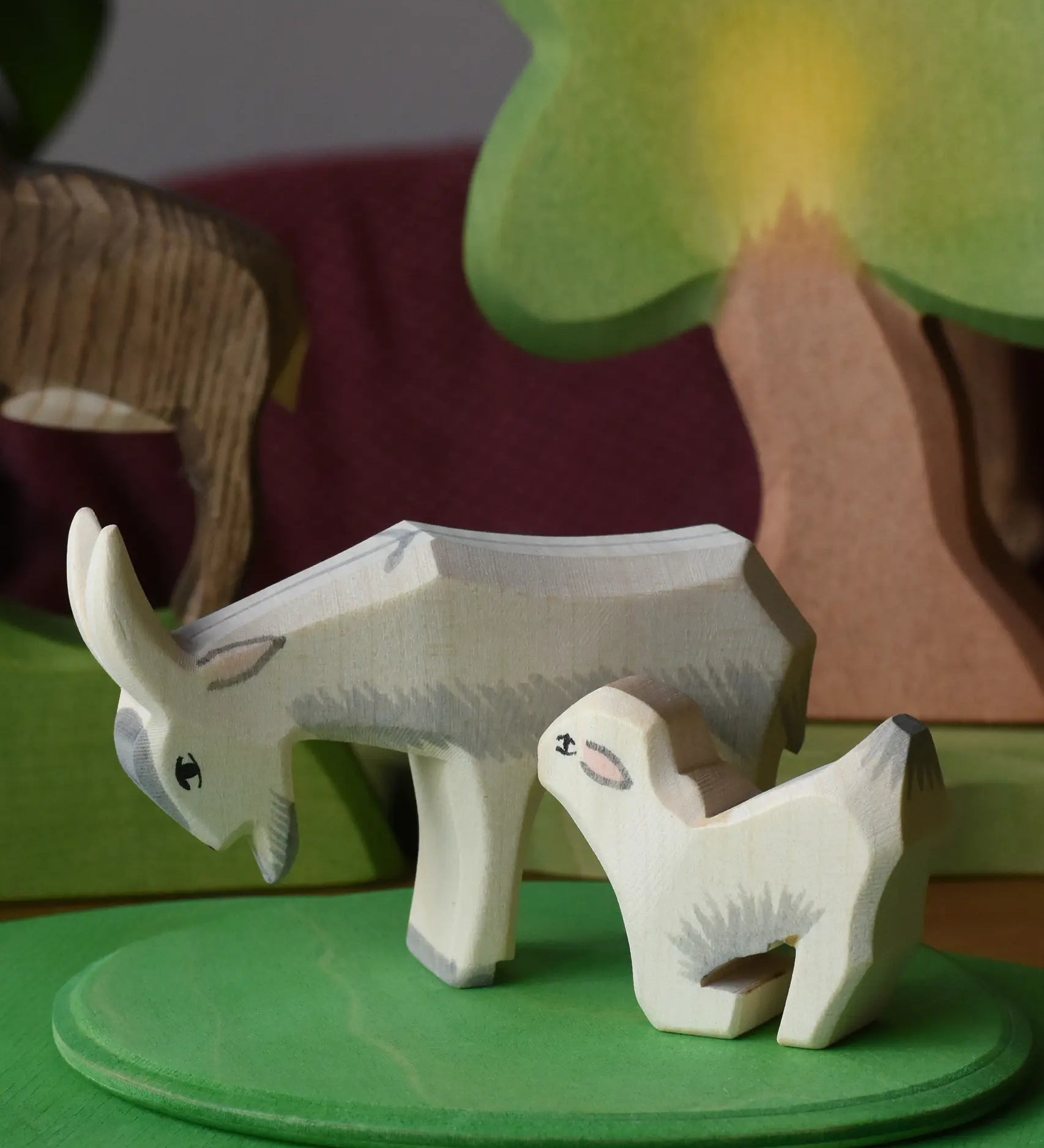 Ostheimer drinking kid wooden toy figure placed next to a white goat on a green meadow place with oak trees in the background.