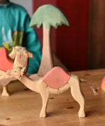 Ostheimer dromedary camel figure with saddle placed in a small world play set up with other camels and a tall palm tree in the background.