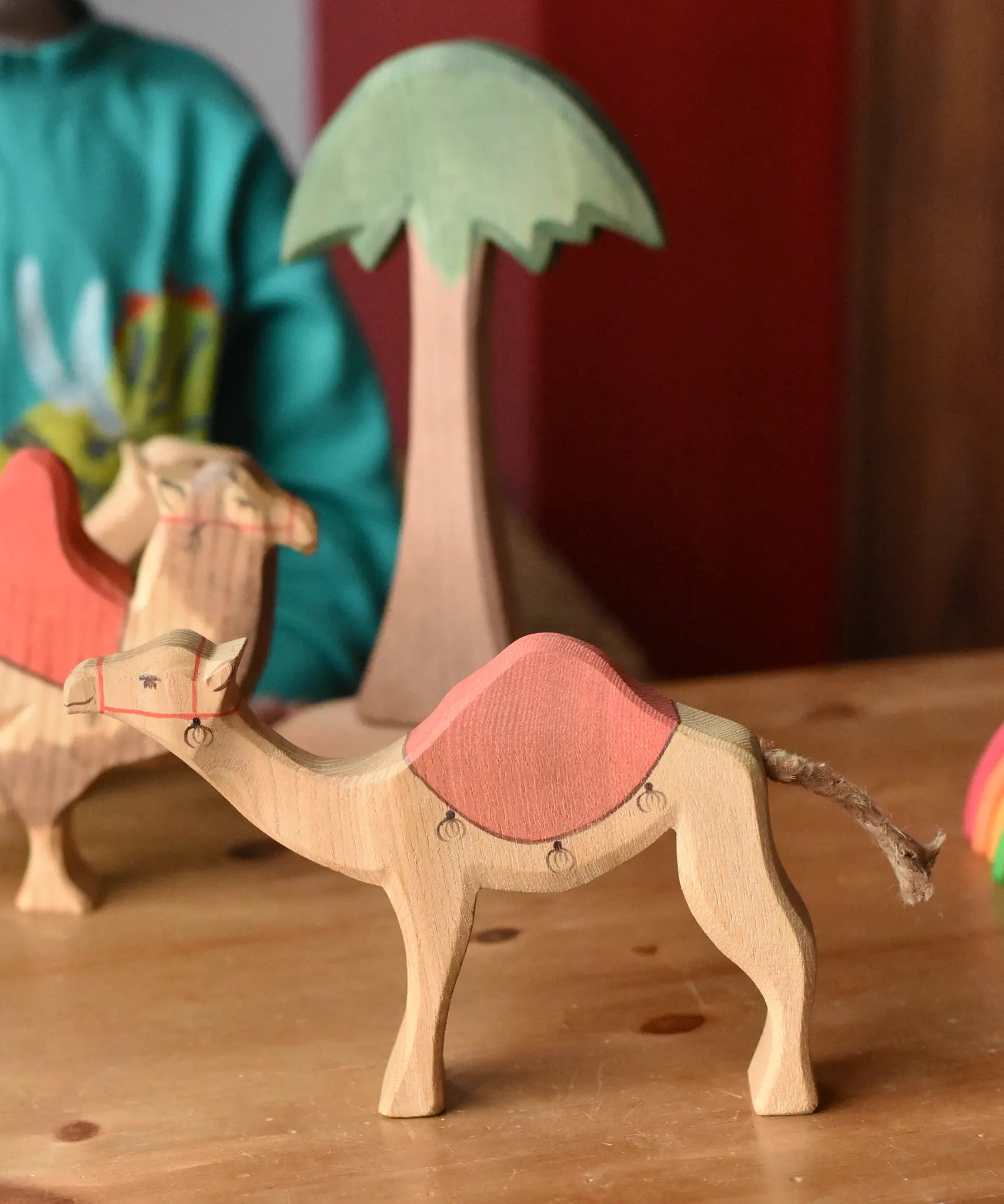Ostheimer dromedary camel figure with saddle placed in a small world play set up with other camels and a tall palm tree in the background.